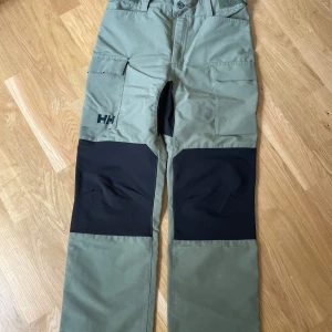 Gröna cargopants från Helly Hansen - Säljer ett par gröna cargopants från Helly Hansen med svarta förstärkningar på knän och bak. Byxorna har stora fickor på sidorna och är perfekta för utomhusaktiviteter. Klassisk cargomodell med normal passform och tåligt material.