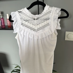 Vit spetsdetaljerad topp från Cubus - Supersöt vit topp från Cubus med spets och volangdetaljer upptill. Ärmlös modell med knappar i nacken och vackert mönstrad spets framtill. Perfekt för en fräsch och feminin look!