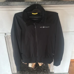 Svart jacka från Sail Racing - Svart jacka från Sail Racing med dragkedja framtill och logga på bröstet. Jackan har hög krage, långa ärmar och ribbade muddar. Perfekt för dig som gillar stilren och sportig design.