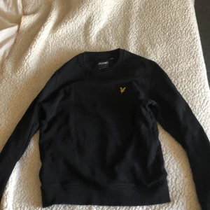 Svart sweatshirt från Lyle & Scott - Säljer en svart sweatshirt från Lyle & Scott med klassisk gul logga på bröstet. Tröjan har rund halsringning och långa ärmar. Perfekt för en stilren och enkel look.