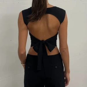 Svart topp med öppen rygg och rosett - Trendig svart topp med breda axelband, fyrkantig halsringning och öppen rygg som knyts med en stor rosett. Aldrig använd!! Perfekt till sommaren, super trendig och populär. Går inte att köpa längre! ❤️
