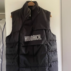 Svart dunväst från Hoodrich - Säljer en svart dunväst från Hoodrich med hög krage och dragkedja. Västen har en stor ficka framtill med Hoodrich-logga i vitt och flera detaljerade sömmar. Perfekt för lager-på-lager-stil.