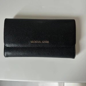 Svart plånbok från Michael Kors - Snygg svart plånbok från Michael Kors i skinn med guldfärgad logga framtill. Plånboken har flera kortfack, fönster för ID och dragkedjefack på baksidan. Perfekt för att hålla ordning på kort och kontanter.