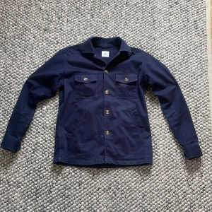 Mörkblå overshirt från John Henric - Säljer en stilren mörkblå overshirt från John Henric med klassisk krage och fem knappar framtill. Två bröstfickor med knappar samt långa ärmar med knapp vid ärmslut. Perfekt för våren och sommar kvällar!