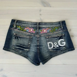 Lågmidjade jeansshorts  - Snygga lågmidjade jeansshorts med broderade blommor i rosa, blått och grönt både fram och bak. Shortsen har slitna detaljer, klassiska fickor och D&G-logga på bakfickan. Perfekta för sommaren! Midjemått: 38cm