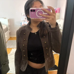 Brun jacka med dekorativa knappar från ONLY - Säljer en brun jacka från ONLY med dekorativa knappar framtill och markerad krage. Jackan har en figurnära passform och är tillverkad i bomull. Perfekt för dig som vill ha en unik och snygg jacka till vardags.