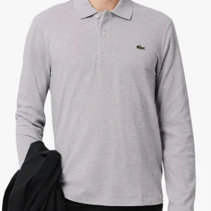 Ljusgrå långärmad pikétröja från Lacoste - Säljer en klassisk ljusgrå långärmad pikétröja från Lacoste med den ikoniska gröna krokodilloggan på bröstet. Tröjan har krage och knäppning med två knappar framtill. Tröjan köptes för 1.450kr