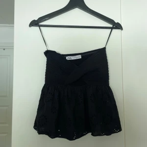 Svart croppad linnetopp från Zara - Säljer en svart croppad linnetopp från Zara med smala axelband och virkad överdel. Nederdelen har broderade blommor och volang. Perfekt för sommardagar och enkel att matcha med jeans eller kjol.