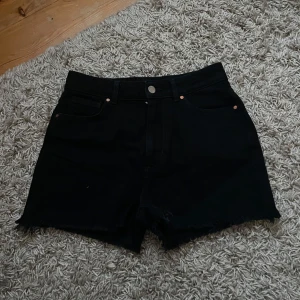 Midwaist denimshorts - Svarta jeansshorts med rå kant och klassisk femficksdesign. Modellen är mid short och har knapp och dragkedja framtill. Perfekta för sommaren och enkla att matcha med olika toppar.
