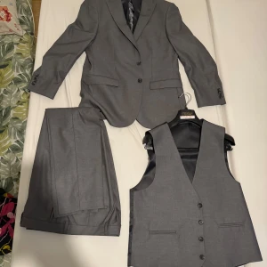 Grå kostym från Paco Romano med väst - Snygg grå tredelad kostym från Paco Romano med kavaj, byxor och matchande väst. Kostymen har slim fit passform och klassisk design med knappar på kavajen och västen. Perfekt för bal eller fest.