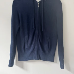 Mörkblå hoodie i kashmir från Soft Goat - Mörkblå hoodie i mjuk kashmir från Soft Goat. Tröjan har dragkedja framtill, huva och två fickor. Perfekt för dig som gillar stilrena och bekväma plagg med premiumkänsla.