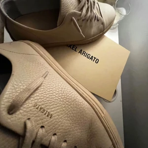 Beige sneakers från Axel Arigato - Säljer ett par snygga och stilrena beigea Axel Arigato-sneakers. Skorna är i princip oanvända – endast testade ett fåtal gånger – så de är i riktigt fint skick. Väldigt sköna och sitter bra på foten, perfekta för både vardag och mer uppklädda outfits.  Designen är minimalistisk och clean, typisk Axel Arigato-stil, vilket gör dem lätta att matcha med det mesta. Ett perfekt val för dig som vill ha kvalitet och stil i ett.  Allt ingår förutom kvitto och dustbag. Nypris är högt, så det här är ett br