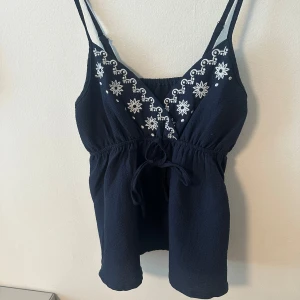Mörkblå linnetopp med broderi - Söt mörkblå linnetopp med smala axelband och vitt broderi framtill. Toppen har knyt i midjan och en lätt v-ringning. Aldrig använd men super fin!
