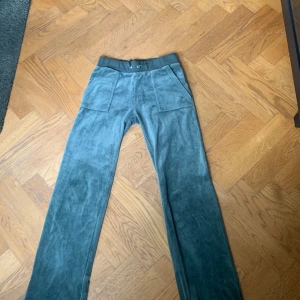Gröna velourbyxor från Juicy Couture - Mysiga gröna velourbyxor från Juicy Couture med bred resår i midjan och raka ben. Byxorna har fickor fram och bak, samt diskret logga broderad på ena fickan. Perfekta för en avslappnad och trendig look.