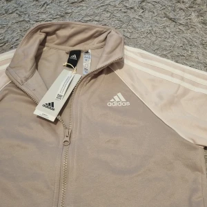 Tröja med zip från Adidas - Säljer en Ny beige/rosa croppad tröja från Adidas med dragkedja och klassiska vita ränder längs ärmarna. Jackan har hög krage och vit Adidas-logga på bröstet. Perfekt för sportiga outfits. Tajt modell.