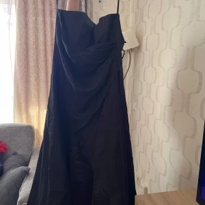 Svart långklänning med smala axelband - Säljer en elegant svart långklänning med smala axelband och draperad detalj framtill. Klänningen har en stilren och klassisk design, perfekt för festliga tillfällen. Materialet är lätt och ger ett fint fall. Nypris 2700kr prislapp finns.