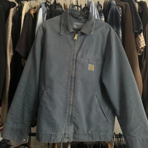 Blå workwear jacka från Carhartt - Säljer en blå workwear jacka från Carhartt med klassisk krage och dragkedja. Jackan har två sidofickor och en bröstficka med Carhartt-logga. Perfekt för dig som gillar streetwear och tidlös stil.