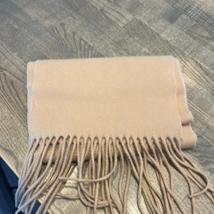 Beige halsduk från Acne Studios - Säljer en klassisk beige halsduk från Acne Studios med fransar i kanterna. Perfekt accessoar för att hålla värmen och lyfta din outfit. Stilren och tidlös design.