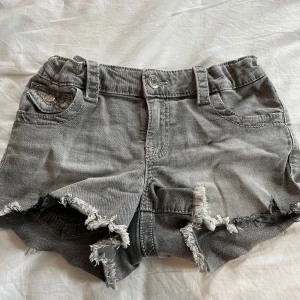 Grå jeansshorts med fransar från Gina Tricot - Snygga grå jeansshorts från Gina Tricot med fransiga benslut och klassisk femficksdesign. Shortsen har bälteshällor och knappgylf. Perfekta för sommaren! Aldrig använts, inga fläckar eller hål 💗
