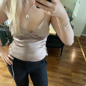 Beige topp med snörning i sidan - Säljer en beige omlottopp med smala axelband och snygg snörning i sidan. Toppen har en v-ringad omlottdesign och är tillverkad i ett glansigt, satinliknande material. Perfekt för att lyfta vilken outfit som helst!