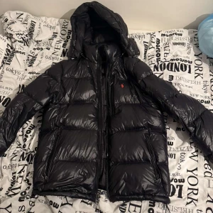 Svart dunjacka från Polo Ralph Lauren - Säljer en svart, glansig dunjacka från Polo Ralph Lauren med huva och dragkedja. Jackan har två fickor framtill och klassisk röd logga på bröstet.    Nypris: 5000kr 