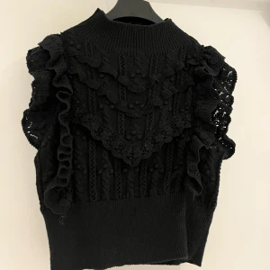 Svart stickad väst med volang från Zara - Säljer en svart stickad väst från Zara med dekorativa volanger och strukturstickat mönster. Hög hals och ribbad nederkant ger en snygg siluett. Perfekt att styla över en skjorta eller t-shirt för en trendig look.