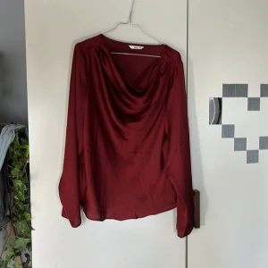 Vinröd blus med draperad hals från Ellos - Säljer en vinröd blus från Ellos med snygg draperad halsringning. Blusen har långa ärmar och är tillverkad i ett glansigt, mjukt material som ger en elegant känsla. Perfekt att styla med jeans eller kjol.