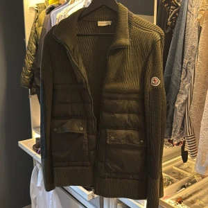 Olivgrön stickad hybridjacka från Moncler - Säljer en olivgrön stickad hybridjacka från Moncler med quiltade partier framtill och två fickor med lock. Jackan har dragkedja, ribbade muddar och Moncler-logga på ärmen. Perfekt för lager-på-lager och snygg till jeans. Modell : Cambales Giubotto