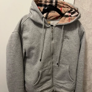  Burberry tröja - Snygg grå hoodie från Burberry med dragkedja och klassiskt rutigt foder i huvan. Jackan har två fickor framtill och justerbar huva med snören. Perfekt för en avslappnad och stilren look. Helt äkta