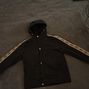 Svart vindjacka med huva från Gucci - Säljer en svart vindjacka från Gucci med huva och tryckknappar. Jackan har beigea ränder med GG-logga längs ärmarna och diskret GG-mönster över hela tyget. Perfekt för dig som gillar streetwear och exklusiva märken.