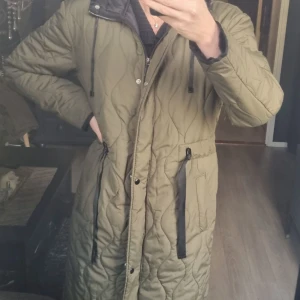 Olivgrön quiltad lång jacka med dragkedja - Säljer en olivgrön lång quiltad jacka med dragkedja och knappar framtill. Jackan har huva med dragsko, svarta detaljer vid fickor och midjebälte. Perfekt för kyligare dagar och har en snygg, avslappnad passform.