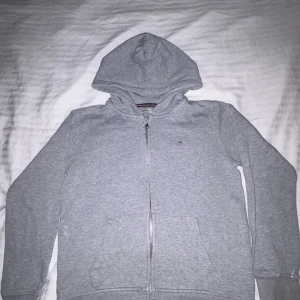 Grå hoodie med dragkedja från GANT - STORLEK 158-164cm.                               Säljer en klassisk grå hoodie från GANT med dragkedja framtill och två fickor. Tröjan har huva och ribbade muddar vid ärmslut och nederkant. Perfekt för om man är laidback. Köpt från KIDSBRANDSTORE.