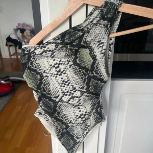 Ormmönstrad baddräkt med one shoulder - Säljer en snygg baddräkt med ormmönster i svart, grått och grönt. Modellen har en axel och hög benskärning för en trendig look. Perfekt för stranden eller poolen! Strl 34 från hm använd en gång 