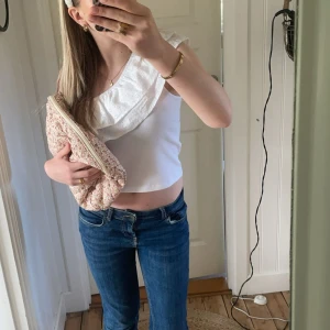 Vit oneshoulder topp med volang - Säljer en vit oneshoulder topp med bred volangdetalj i broderad spets. Toppen är ribbad och har en smal axelrem på ena sidan. Perfekt för sommardagar och enkel att matcha med jeans eller kjol.