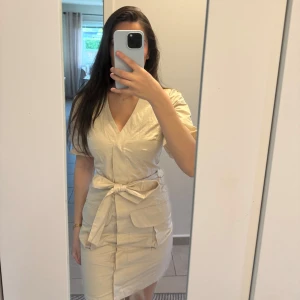 Beige cargoklänning nakd  - Säljer en beige omlottklänning från Sara Sieppi x NA-KD i storlek 34. Klänningen har v-ringning, knytband i midjan, stora fickor framtill och kort ärm. 