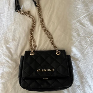 Svart quiltad axelväska från Valentino - Säljer en svart quiltad axelväska från Valentino med guldfärgade kedjor och detaljer. Väskan har ett lock med vridlås och logga framtill. 