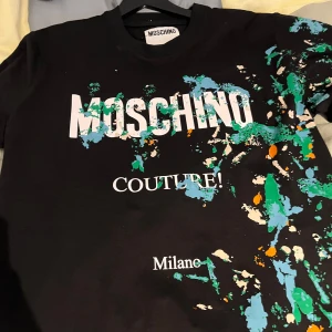 En svart t shirt från Moschino - En svart t shirt från Moschino med färgstänk den är mycket fin och unik perfekt för dig som vill ha något fint och exklusivt samt unikt