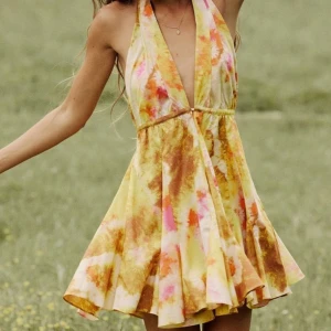 Färgglad omlottklänning med halterneck - Säljer en kort omlottklänning med halterneck och djup v-ringning. Klänningen har ett färgstarkt batikmönster i gult, orange och rosa toner. Perfekt för varma dagar och strandhäng. Kjolen är utställd och ger en luftig känsla.
