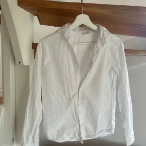 Vit skjorta från Kappahl - Klassisk vit skjorta från Kappahl med krage och knäppning framtill. Skjortan har lång ärm och är tillverkad i ett lätt bomullstyg. Perfekt för en stilren look.