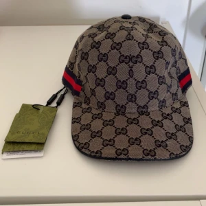 Gucci keps med GG-mönster - Snygg keps från Gucci i beige med klassiskt GG-mönster över hela kepsen. Har röda och blå ränder på sidorna och justerbar rem baktill i svart läder. Märkeslapp och etikett finns kvar.
