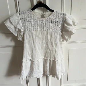 Vit blus med volangärmar och spetsdetaljer - Säljer en söt vit blus med volangärmar och spets upptill. Blusen har en lös passform och dekorativa detaljer. Perfekt för vår och sommar! Sparsamt använd 