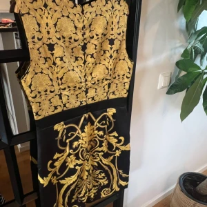 Svart och guldmönstrad klänning från Moschino - Säljer en svart klänning från Moschino med iögonfallande barockinspirerat guldmönster. Klänningen är ärmlös, har rund halsringning och figurnära passform. Perfekt för dig som vill sticka ut med en lyxig och elegant look.på klänningen står d att d är storlek xxl men upplever den som storlek 38
