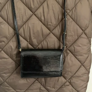 Svart axelväska från H&M - Snygg svart axelväska från H&M i glansigt material med struktur. Väskan har justerbart axelband och stängs med lock. Perfekt för att bära det viktigaste till vardags eller när du vill ha en stilren look.