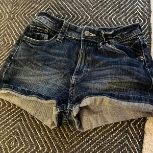 Mörkblå jeansshorts från H&M - Snygga mörkblå jeansshorts från H&M med klassisk femficksdesign och uppvikta benslut. Shortsen har normal passform och detaljerade sömmar samt knappstängning fram. Perfekta för varma dagar!