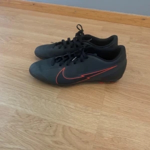 Svarta Nike fotbollsskor med röda detaljer - Säljer ett par svarta Nike fotbollsskor med röda detaljer och snörning. Skorna har rund tå och platt sula med fasta dobbar för bra grepp på planen. Perfekta för dig som vill ha stilrena och funktionella skor till match eller träning. OBS Box medföljer ej 