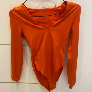 Orange/röd långärmad bodysuit med v-ringning - Säljer en snygg orange/röd bodysuit med lång ärm och djup v-ringning. Perfekt för att styla med jeans eller kjol för en trendig look. Mjukt och stretchigt material som sitter bekvämt på kroppen.