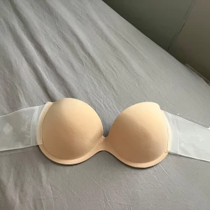 Beige axelbandslös bh med transparenta band - Säljer en beige axelbandslös bh med transparenta band och bygel. Perfekt när du vill ha en osynlig look under kläderna. Medföljer extra spänne för justering framtill