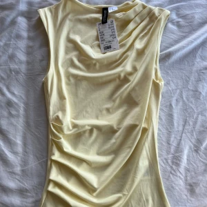 Ljusgul ärmlös topp från Divided - Säljer en ljusgul ärmlös topp från Divided by H&M. Toppen har draperade detaljer vid axeln och en mjuk, stretchig känsla. Perfekt för att addera lite färg till din garderob! Har aldrig använts 💓