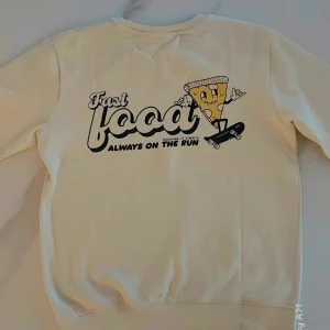 Beige sweatshirt med pizzatryck - Säljer en beige sweatshirt med tryck av en tecknad pizzaslice på skateboard och texten 'Fast food always on the run' både fram och bak. Tröjan har rund hals och långa ärmar. Perfekt för dig som gillar roliga prints!              Det finns en liten defekt på höger ärm. Det finns en viss svärta.  