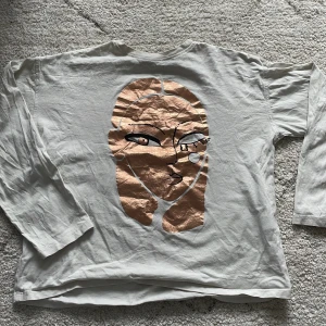 Vit långärmad tröja med ansiktsmotiv från Zara - Vit långärmad tröja från Zara med ett stort, stiliserat ansikte i metallic-rosa och svarta detaljer på framsidan. Tröjan har en avslappnad passform och är gjord i mjukt bomullsmaterial. Perfekt för dig som gillar unika prints!